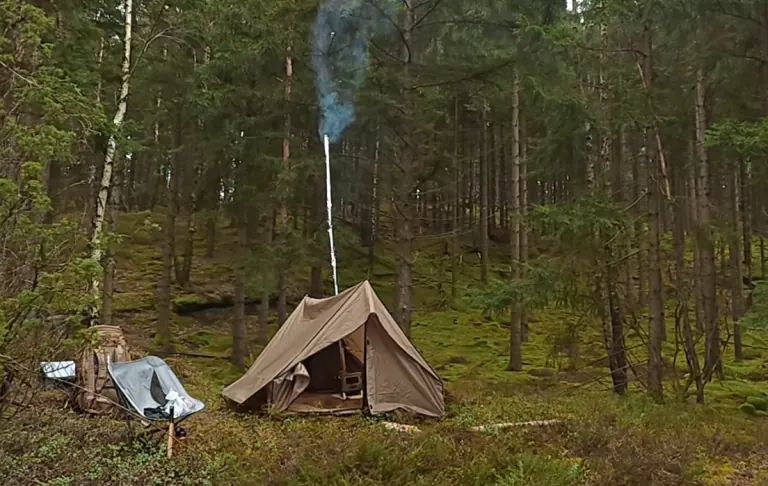 Karstā telts ar plīti mežā, dūmi paceļas no skursteņa, koku ieskauta. Pie telts atrodas kempinga krēsls un mugursoma.