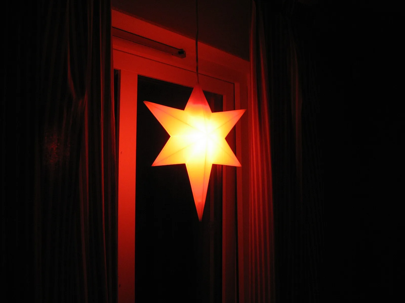 En tänd röd-orange adventsstjärna av papper hänger i ett mörkt fönster med röda gardiner