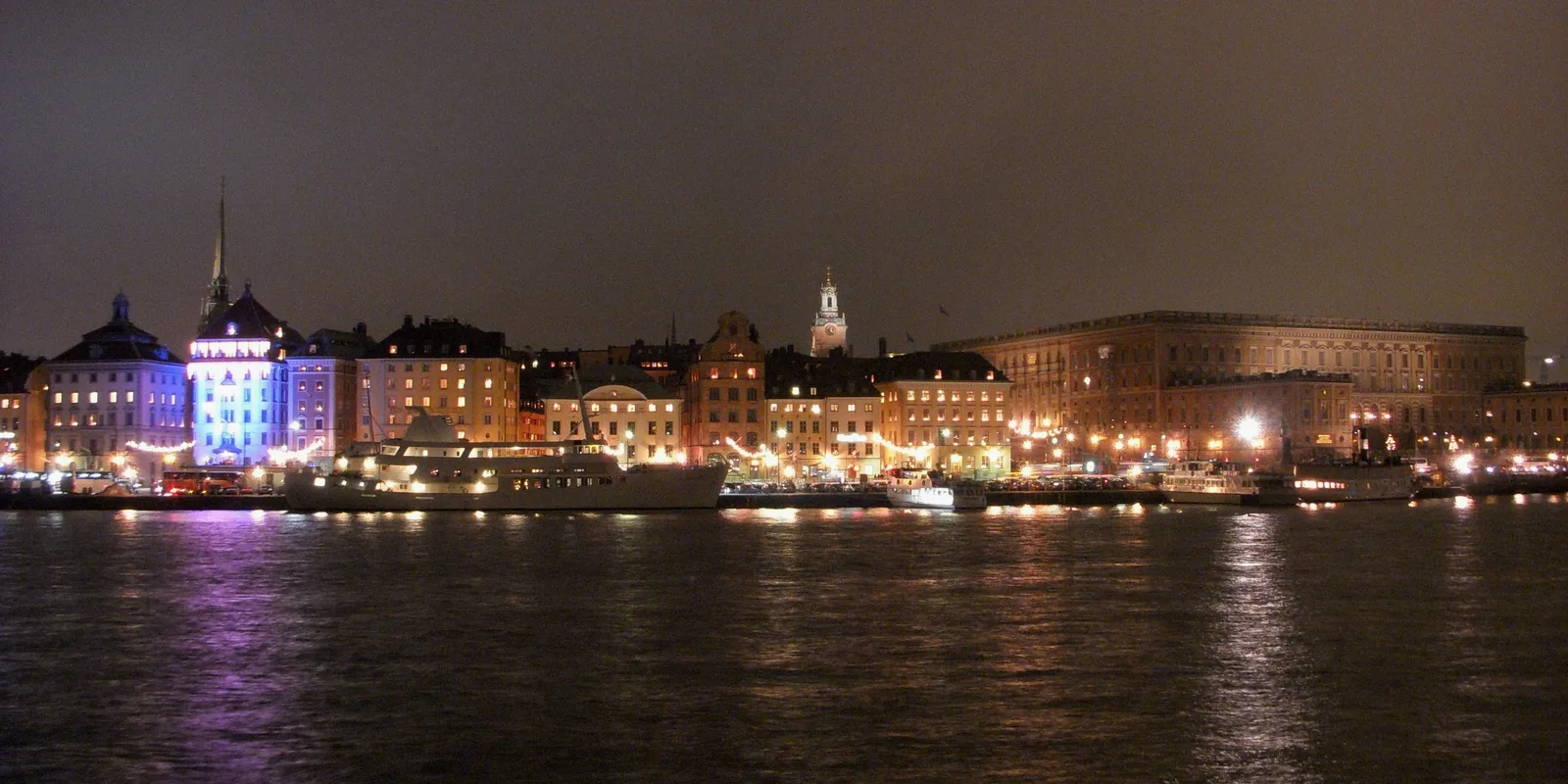 Gamla stan och Stockholms slott i kvällsljus med julbelysning, sett över vattnet med båtar vid kajen.