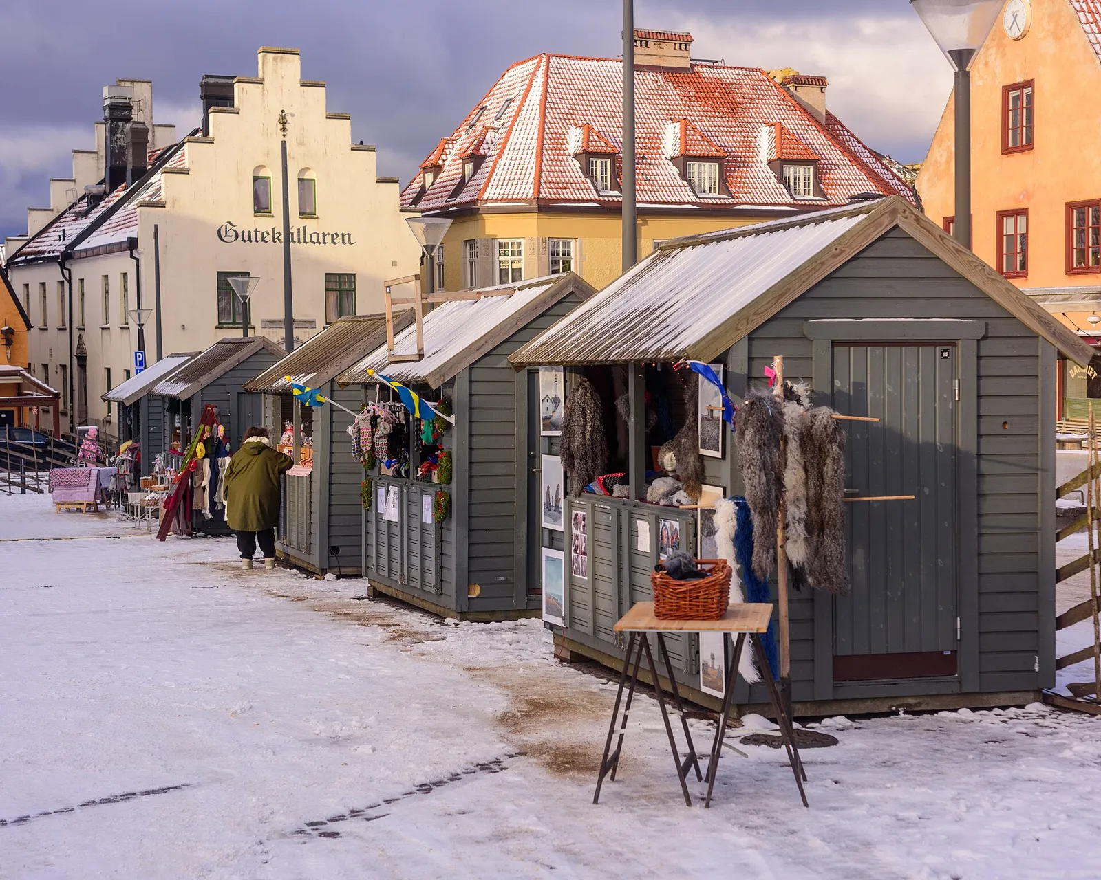 Små gråmålade julmarknadsstånd i trä på ett snöigt torg framför gamla gavelhus i Visby i skymningen.