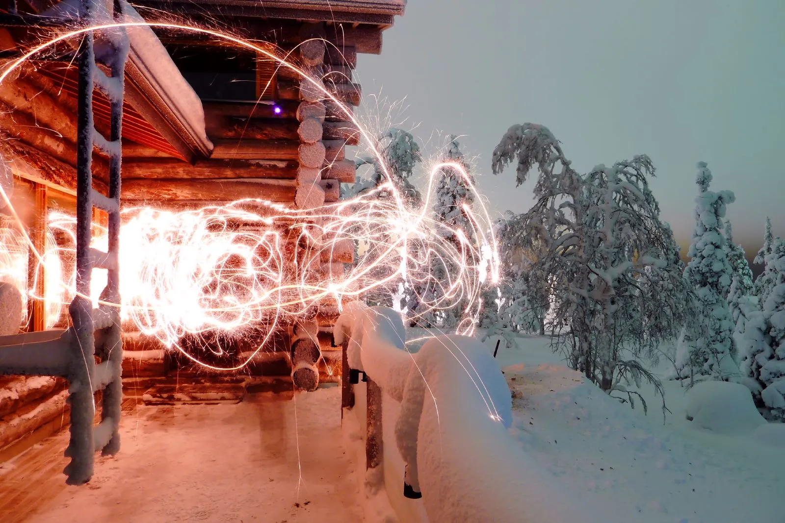 Tomtebloss ritar ljusslingor framför en snötäckt timmerstuga på fjället en vinterkväll, med frostiga granar bredvid.