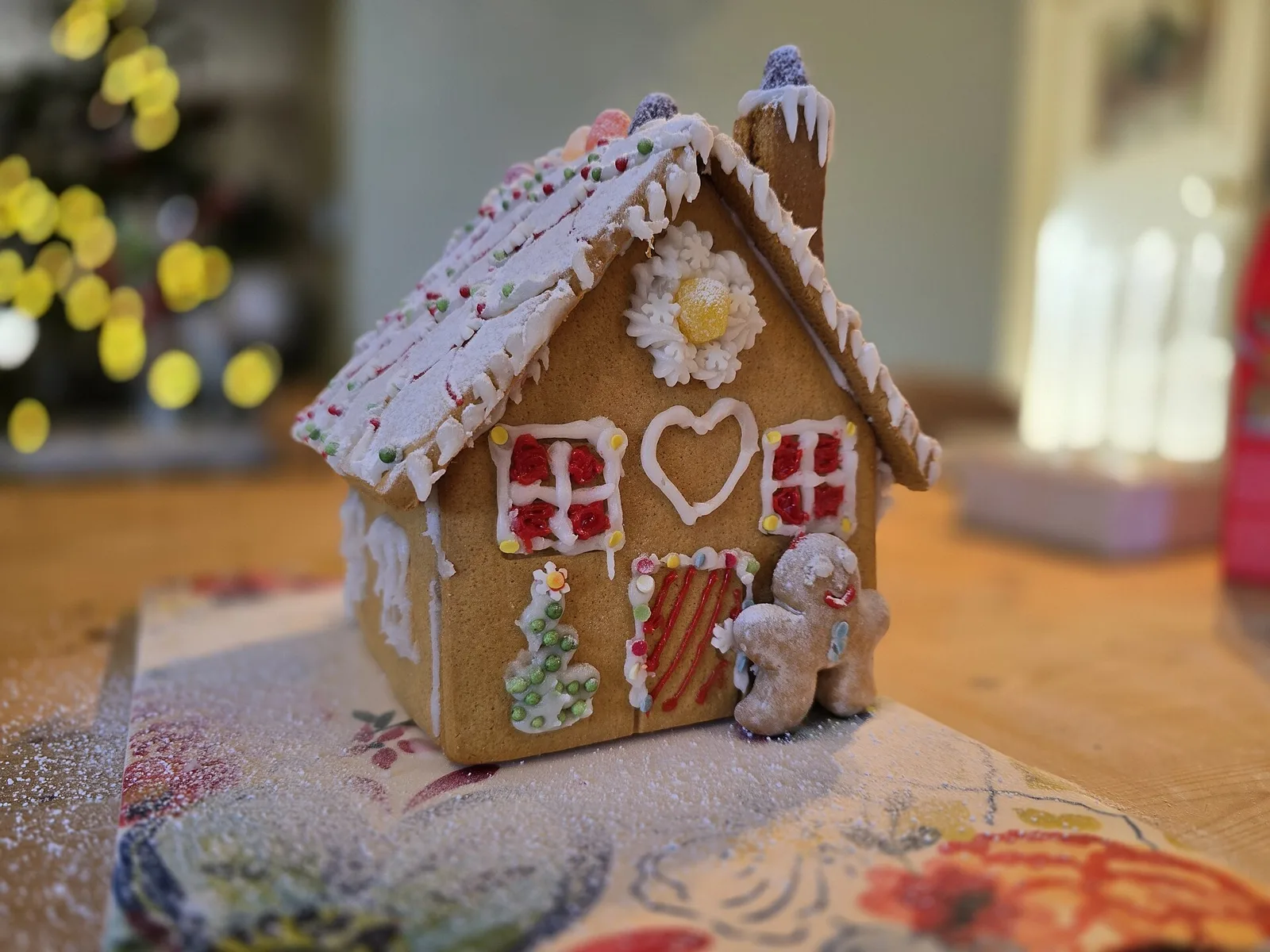 Ett litet pepparkakshus med vit kristyr som snö på taket, röda fönster och en pepparkaksgubbe framför, på ett bord med julbelysning i bakgrunden