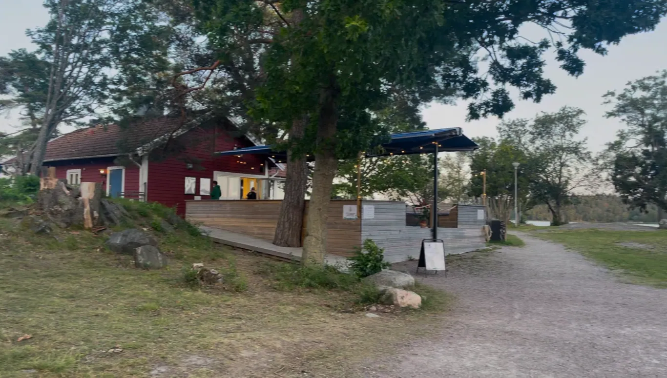En rød hytte med udendørs siddepladser og grønne områder omkring det. En vej fører op til det.