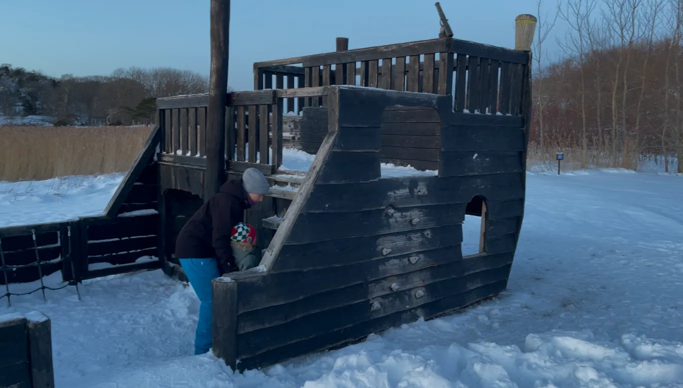 Barn leker på en lekeplass i snøen, klatrer en svart trestruktur formet som et skip.