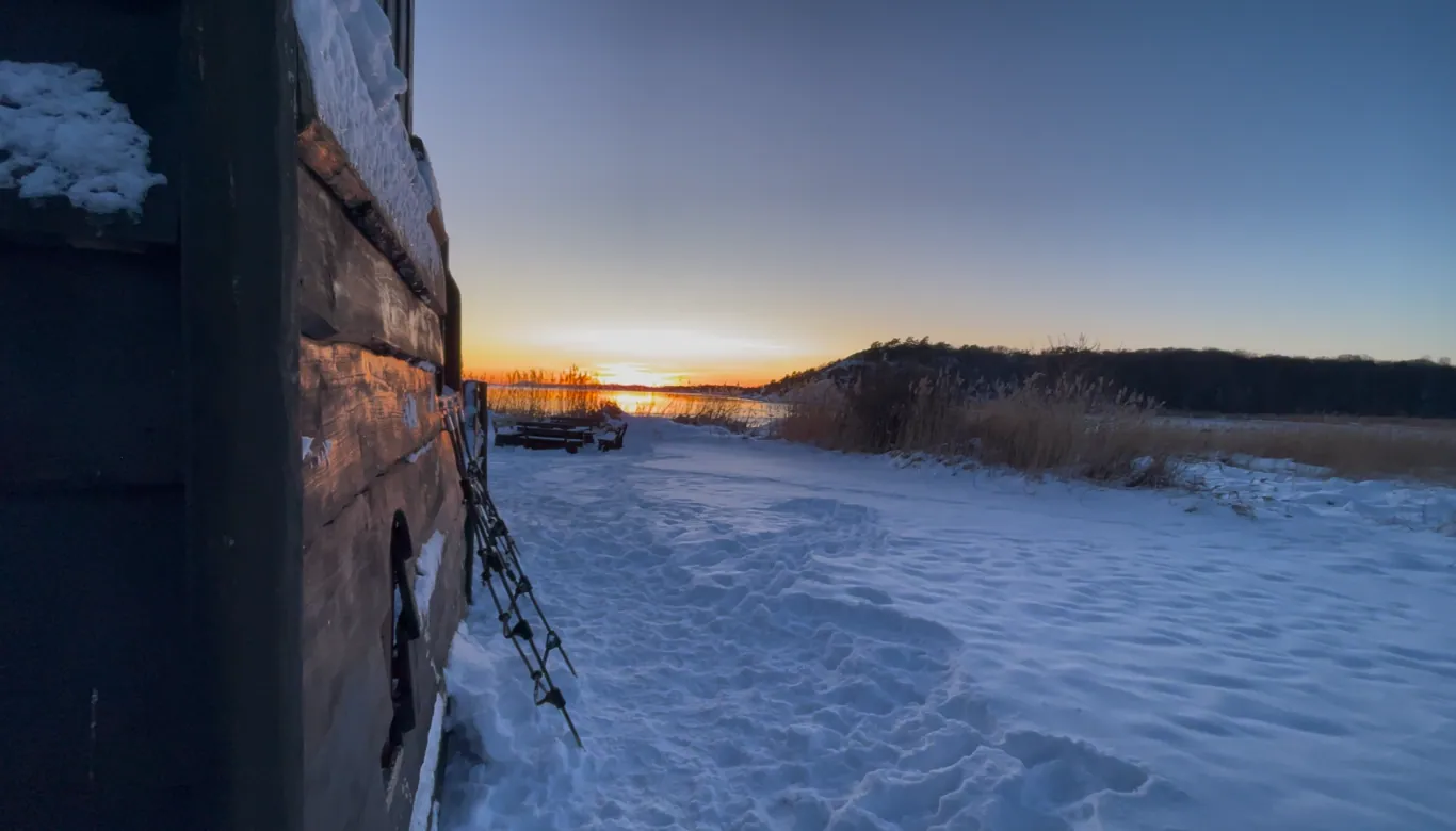 Vinterlandskap ved vannet med snø, en trestruktur i forgrunnen og en solnedgang i bakgrunnen.
