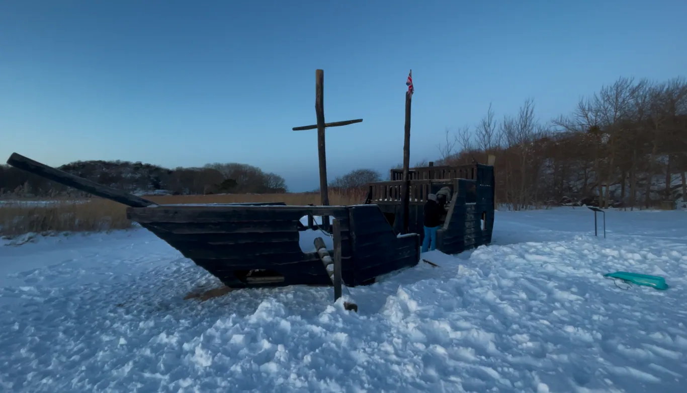 En gammel båt hviler på snødekt grunn med et kors på masten og trær i bakgrunnen.