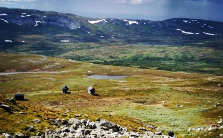 Stort bjerglandskab med slidte grønne enge og små puljer af vand. Sne på bjergtoppe.