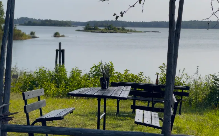 En utendørs sittegruppe ved en innsjø med benker og bord, omgitt av grøntområder og en øy i bakgrunnen.