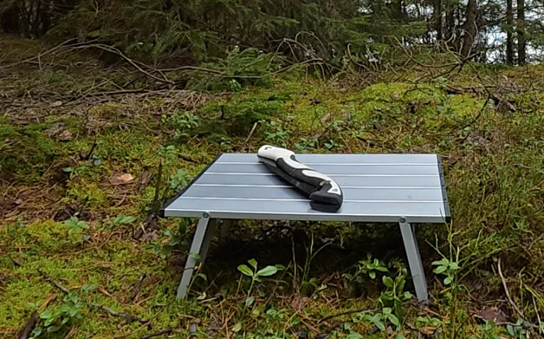 Kompaktiškas kempingo stalas miške su ilgu juodu objektu ant paviršiaus.