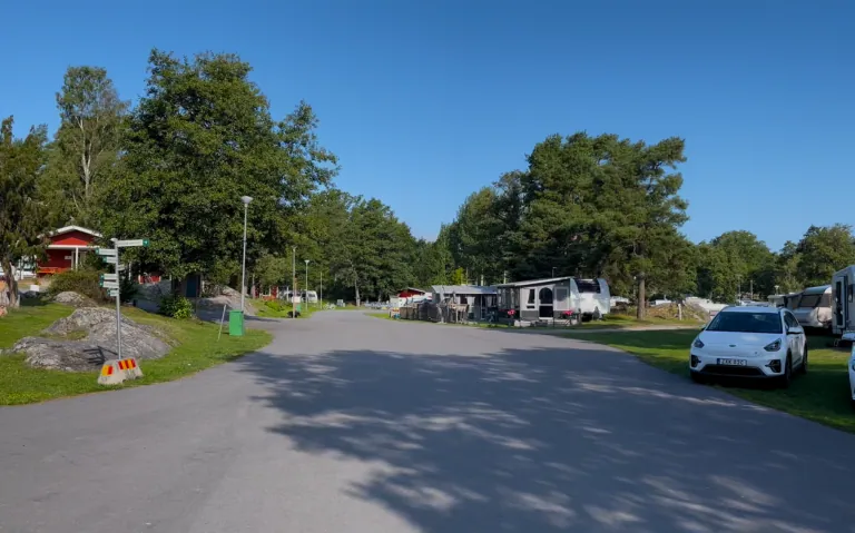 En campingplass med vei, trær og campingvogner under en klar blå himmel.