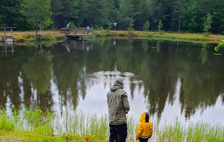 Twee mensen bij een rustig meer omringd door bomen, een volwassene en een kind in gele regenkleding.