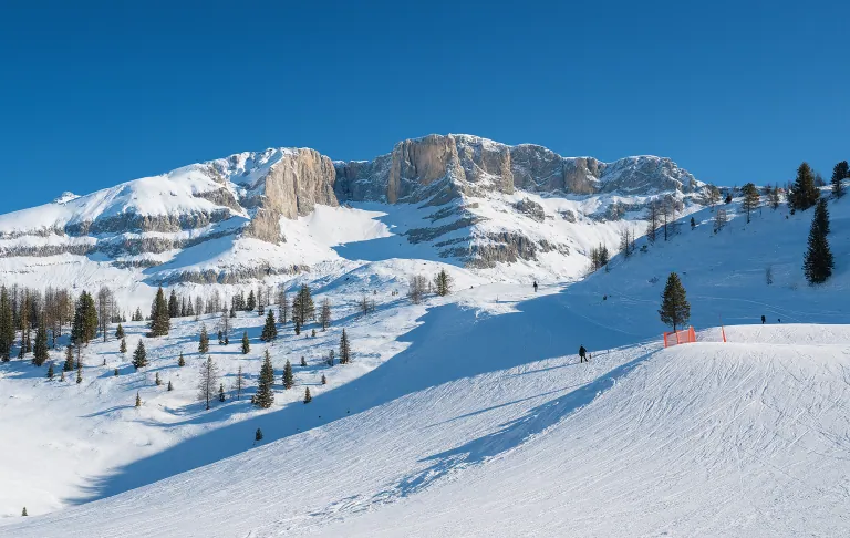 Abbildung von schneebedeckten Bergen und Tälern unter einem klaren blauen Himmel, mit Skifahrern im Hintergrund.