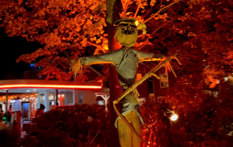 Scarecrow līdzīga skulptūra oranžā apgaismojumā, rudens koku ieskauta.