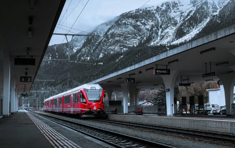 Et rødt tog på en snedækket station med bjerge i baggrunden.