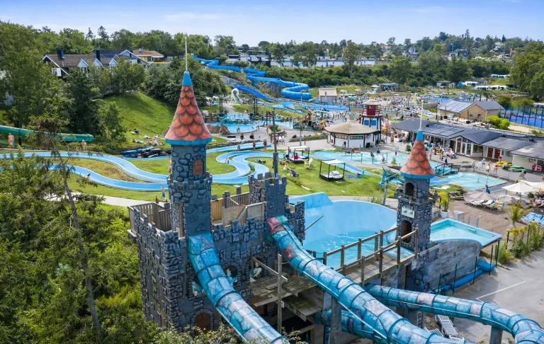 Een waterpark met lange glijbanen, een kasteel en ligstoelen in een groene omgeving.