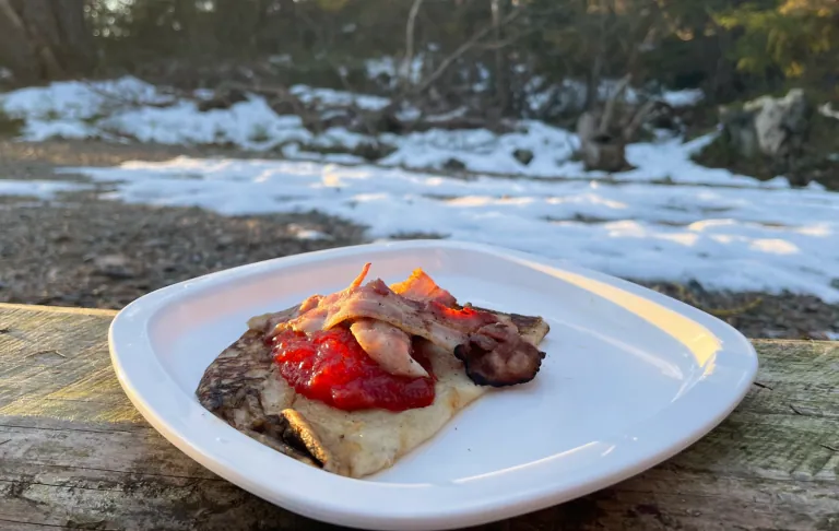 Tallerken med en pandekage toppet med jordbærsyltetøj og bacon, omgivet af sne og natur.
