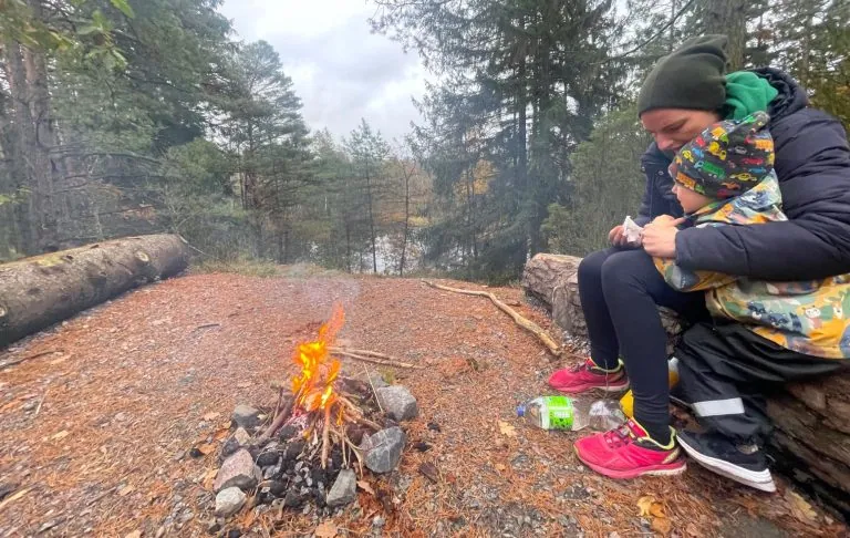 En person sidder ved et lejrbål i skoven, omgivet af træer og en sø.