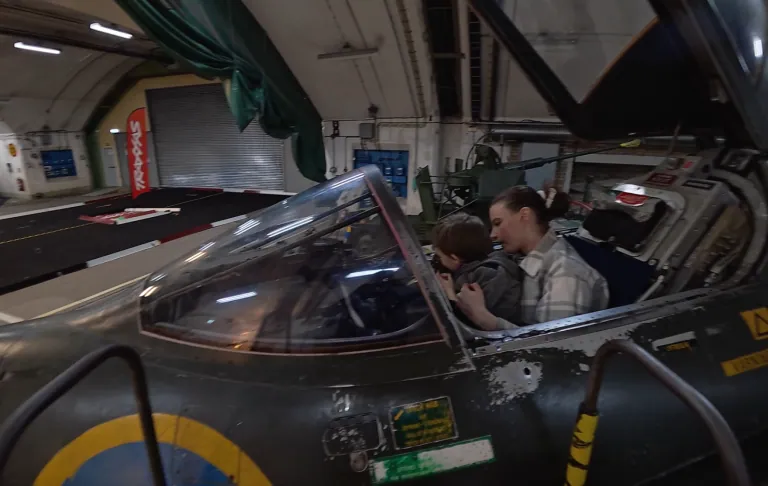 Två personer sitter i cockpit på ett militärt flygplan inomhus.