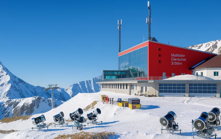 Illustration av Skidstation med röda fasader, snökanoner och berg i bakgrunden. Klarblå himmel.