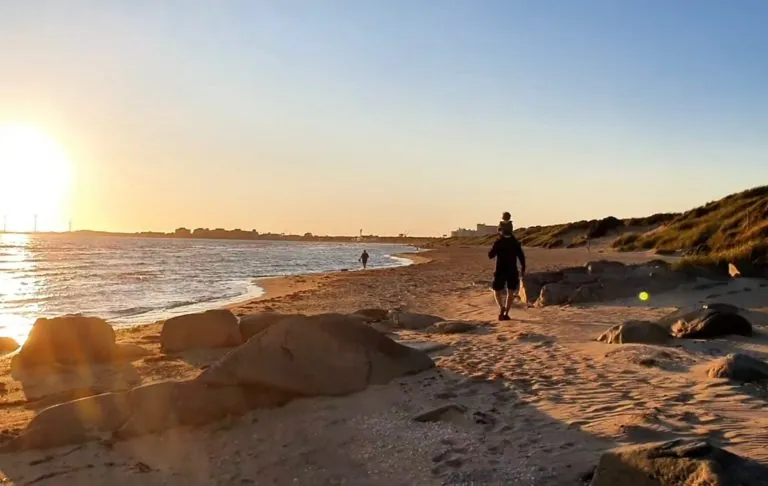 Person går längs en strand vid solnedgången, med vatten och klippor i bakgrunden.