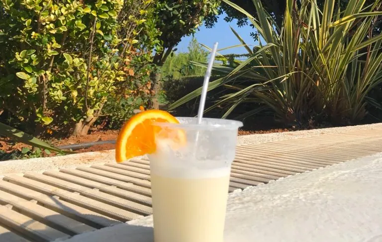 En plastmugg med vit drink och en skiva apelsin, placerad på en grå yta.