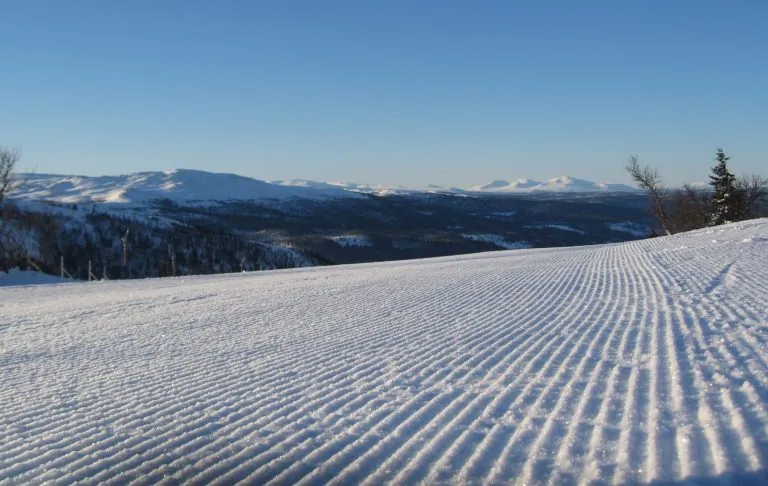 Lumipeitteinen maisema, jossa on jälkiä lumessa ja vuoria taustalla kirkkaan sinisen taivaan alla.