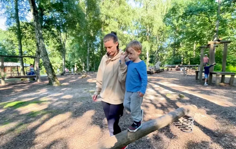 A woman walks with a child on a balance beam in a playground surrounded by trees.