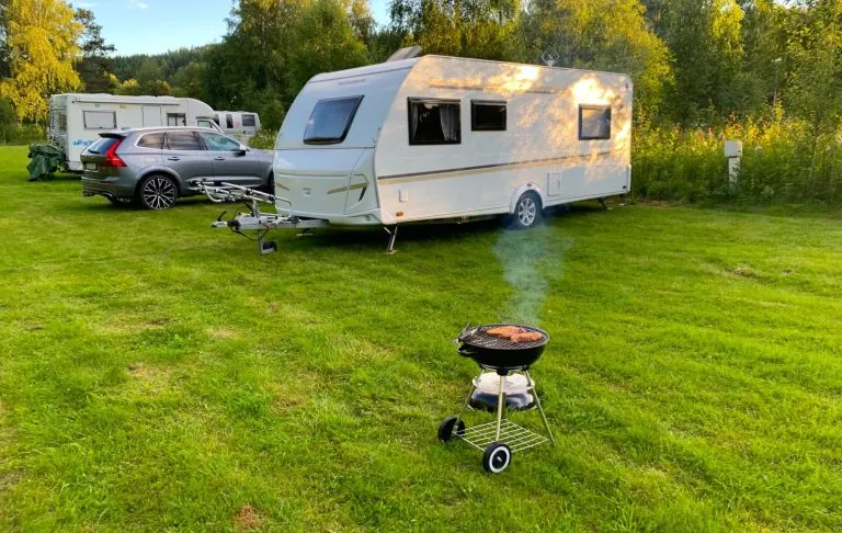 A caravan on a lawn with a smoking grill in the foreground.