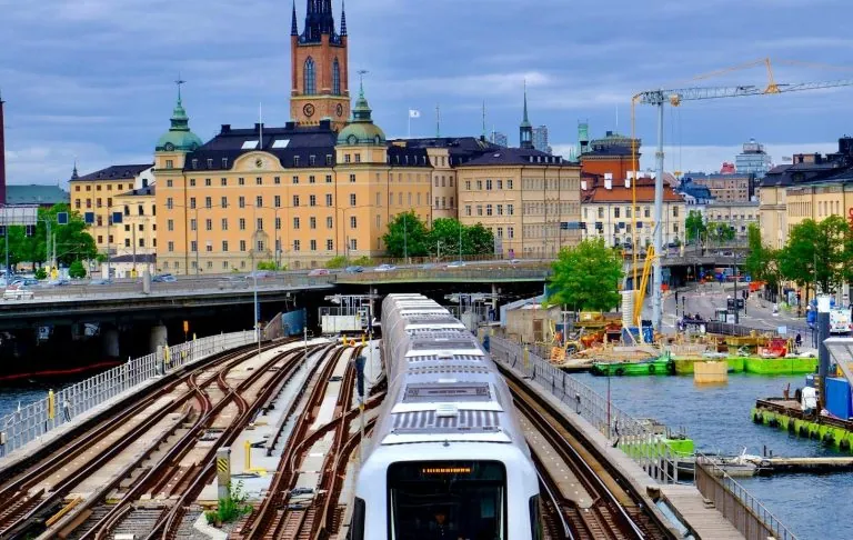 Ein Zug passiert eine Stadt mit Gebäuden und Wasser im Hintergrund.