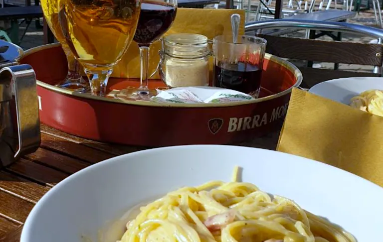 Tallerken med pasta, to drinks og en dessert i baggrunden på en udendørs terrasse.