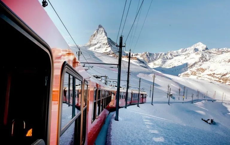 Et tog passerer gennem snedækket landskab med Matterhorn og bjerge i baggrunden.