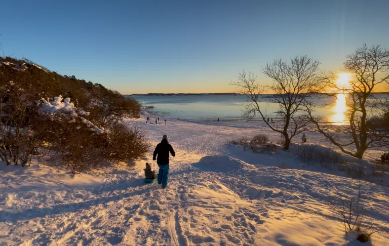 En person går ned en snødekt strand ved solnedgang over havet.