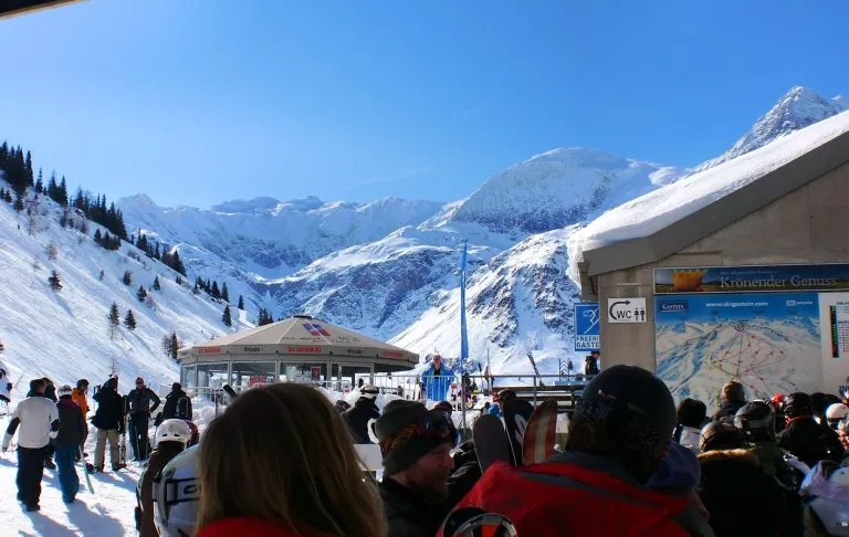 Menschen in einem Skigebiet mit schneebedeckten Bergen und einem klaren blauen Himmel im Hintergrund.