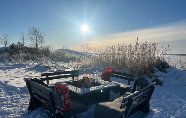 En vinterpiknik med snødekte benker og bord, solstråler i bakgrunnen.