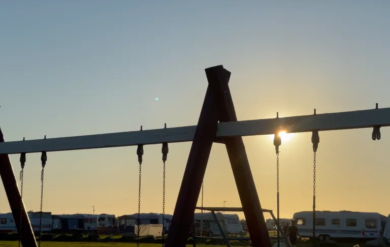 Silhouet van een speelstructuur met zonsondergang op de achtergrond en kampeerders in de verte.