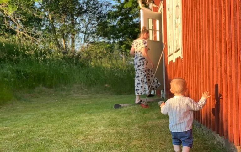 Valges kampsunis laps kõnnib täiskasvanu poole, kes töötab päikesepaistelise ilmaga punase suvila juures.