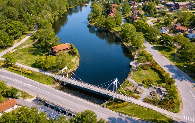 Skats uz ezeru šķērsojošu tiltu, ko ieskauj apstādījumi un mājas.