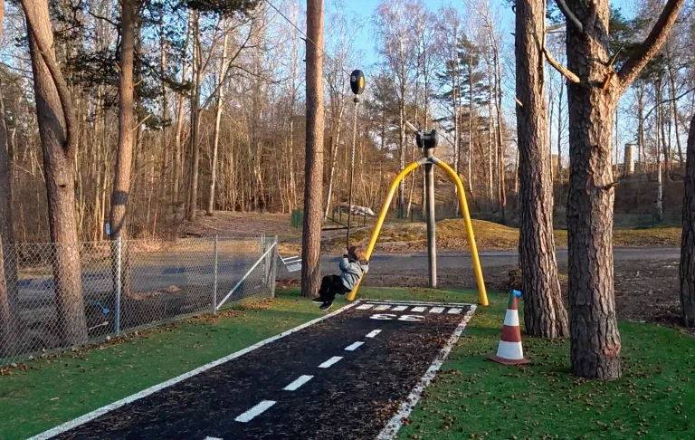 En gungställning vid en lekplats omgiven av träd och en grusväg.