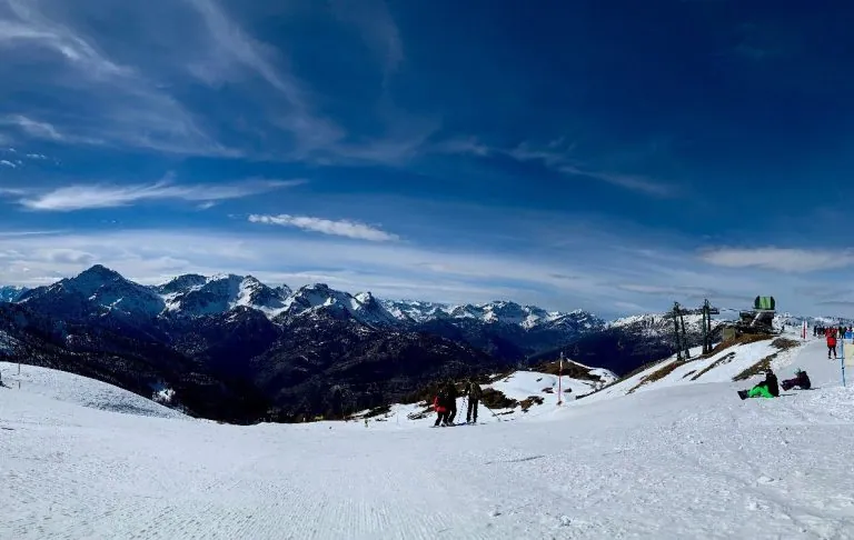 Winterlandschaft mit verschneiten Alpenbergen und einem klaren blauen Himmel, Skifahrer im Hintergrund.
