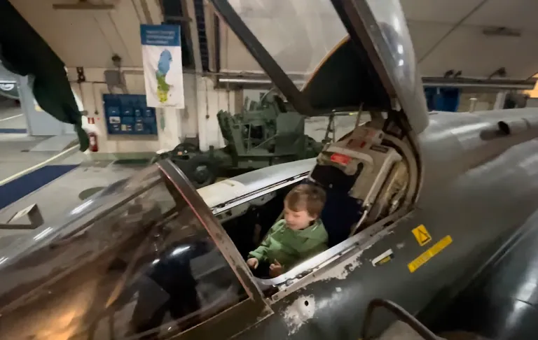Et barn sidder i cockpittet på et militærfly inde i en hangar.