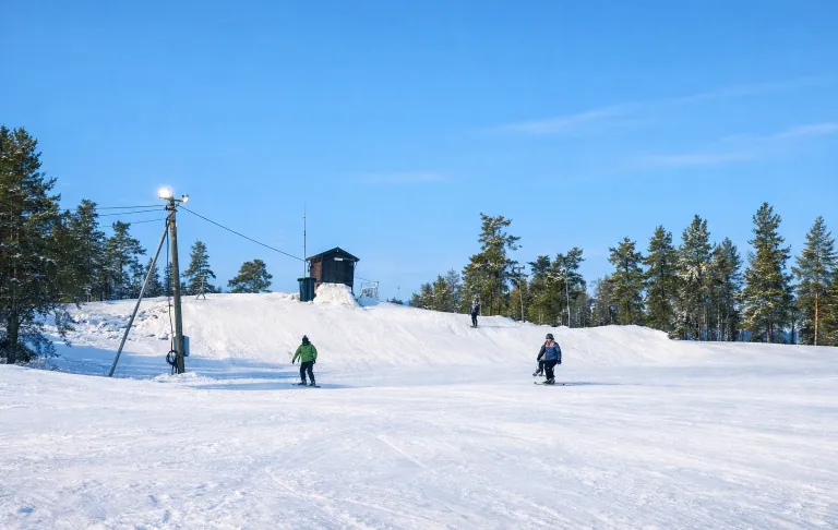 Två personer åker skidor på snöklädd backe med ett hus och träd i bakgrunden.