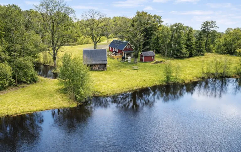 Landligt landskab med røde bygninger ved en sø, omgivet af grønne områder og træer.