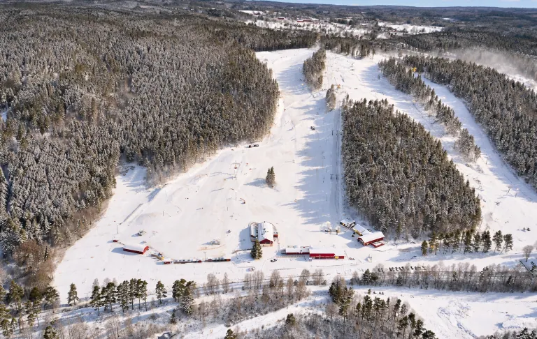 Snötäckt skidbacke med röda byggnader omgiven av täta granar.