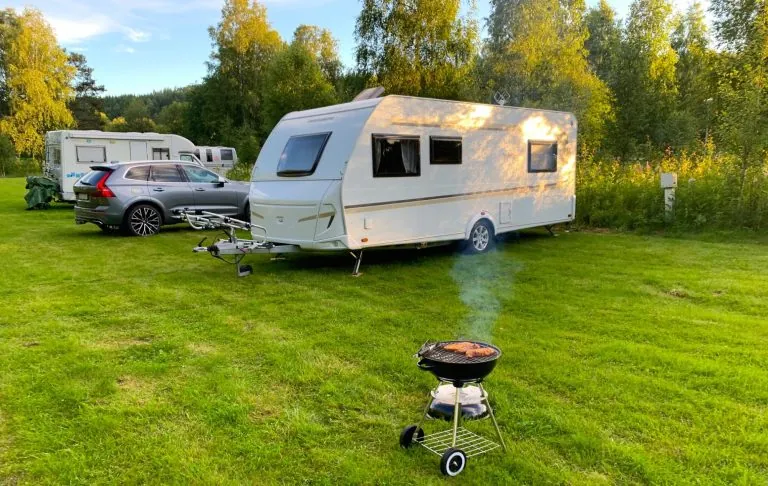 White Caravan auf einem Campingplatz mit einem Grill im Grünen. Der Sonnenuntergang scheint über der Gegend.