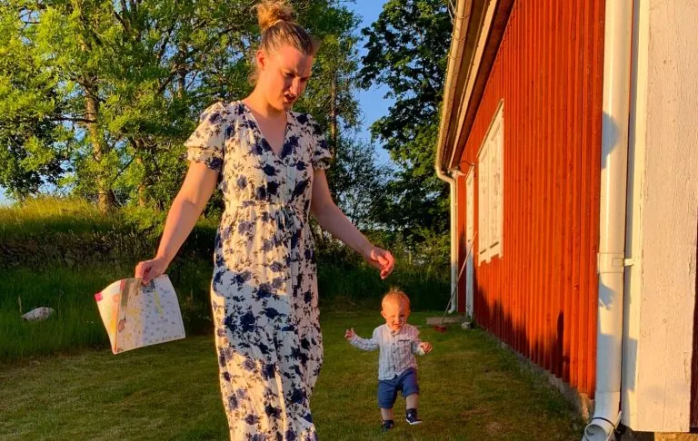 En kvinde i kjole går med et barn udenfor en rød hytte i solskin.