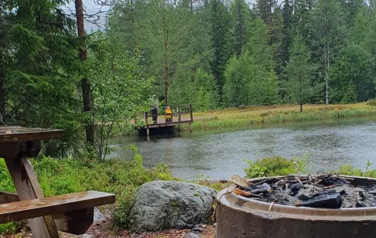 En sø med en dock i skoven, en bænk dæmning og en pejs i forgrunden. Et menneske, der fisker.