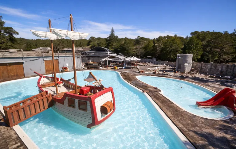 Een kinderbad met bootspel en een glijbaan, omgeven door groen onder een heldere blauwe lucht.