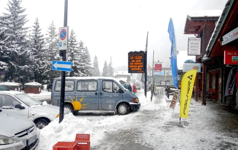 Ziemas ainava ar sniegu, stāvošām automašīnām un veikaliem slēpošanas kūrortā.
