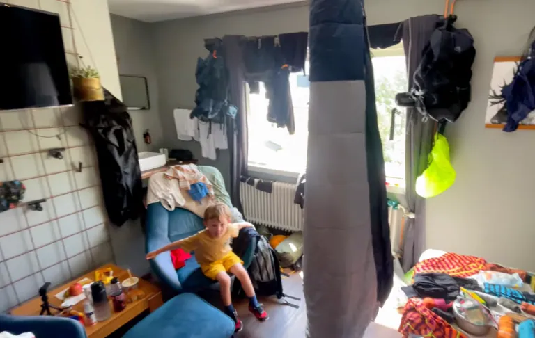 Et barn sidder på en sofa i et rodet rum med tøj og genstande spredt rundt.