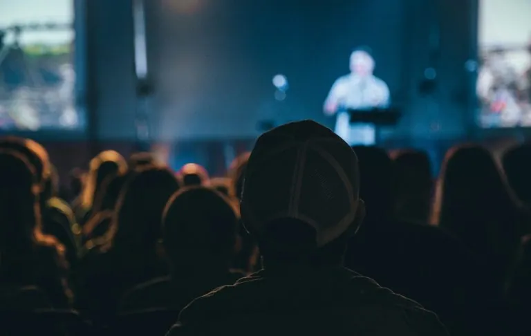 Cilvēki auditorijā skatās runātāju uz skatuves vājā apgaismojumā.